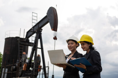Two female engineers stand beside working oil pumps with a white sky background.