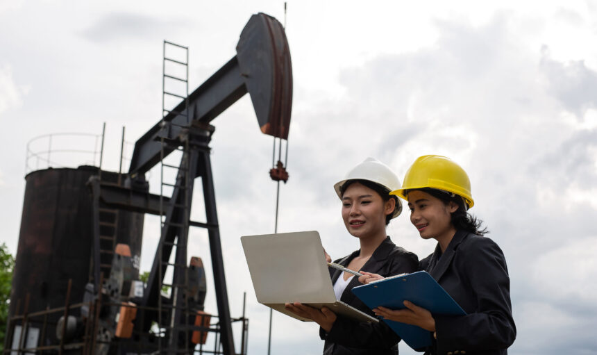 Two female engineers stand beside working oil pumps with a white sky background.